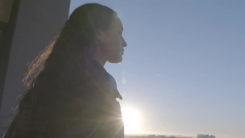 A young woman with long hair standing on a parkinggarage balcony gazing out at the city skyline as the sun sets in warm colors