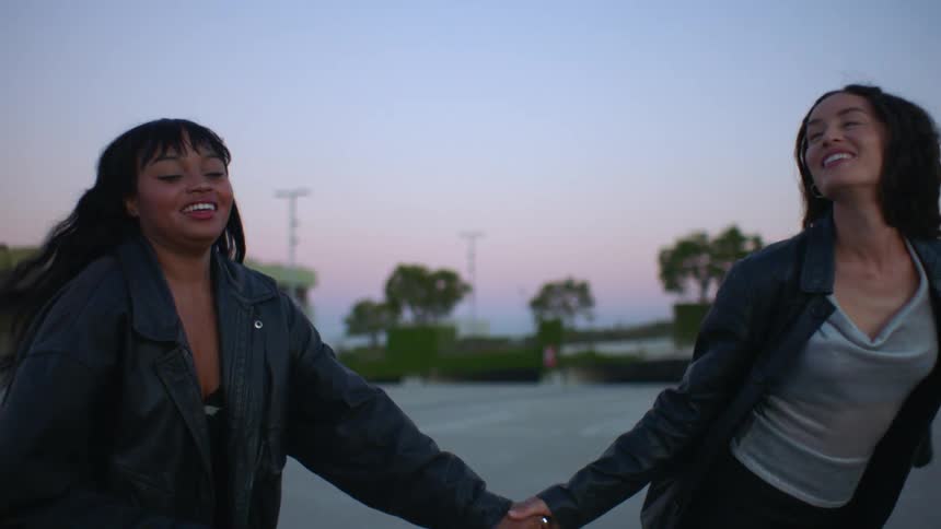 Two female friends holding hands while walking together outdoors in a city at sunset