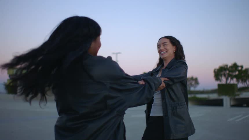 Two women wearing black jackets laugh and smile while holding each others arms and spinning in circles on an open rooftop parking lot during twilight