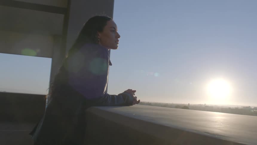 A woman with long dark hair leans against a balcony ledge while looking out at a city during sunset the scene transitions from a wide profile view to a closer shot of her face as she gazes toward the bright horizon