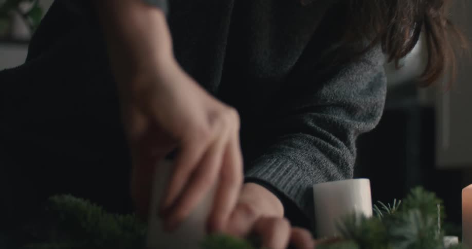 A woman with long dark hair wearing a charcoal sweater focuses intently as she uses both hands to press a thick white candle into a handmade pine wreath