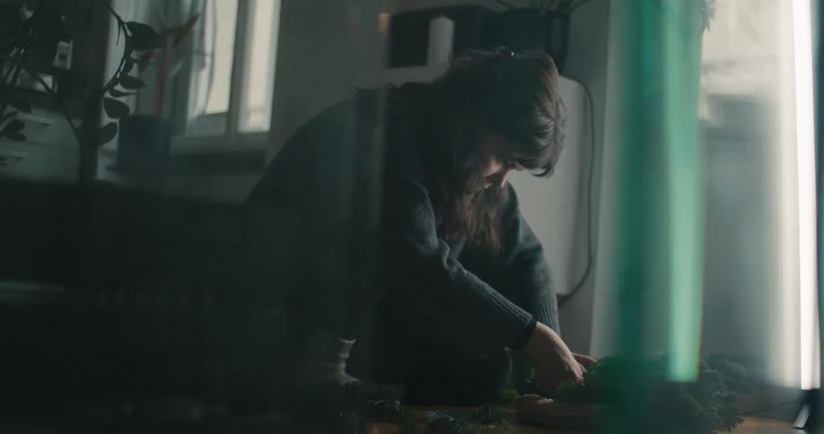 A woman sits on a wooden floor in a dimly lit room carefully arranging evergreen branches into a circular wreath