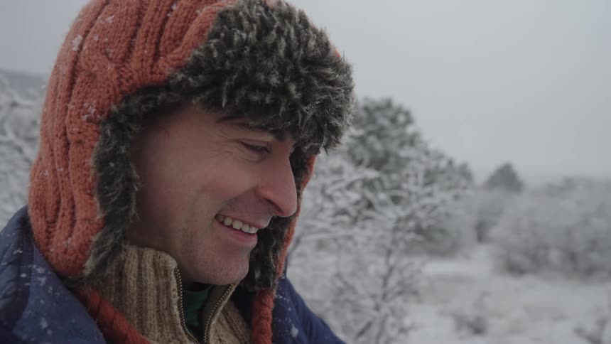 A smiling man wearing an orange and faux fur trapper hat and a knit sweater is outdoors in a snowy landscape