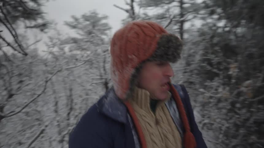 A man in a trapper hat and sweater is laughing and walking through a snowy forested area