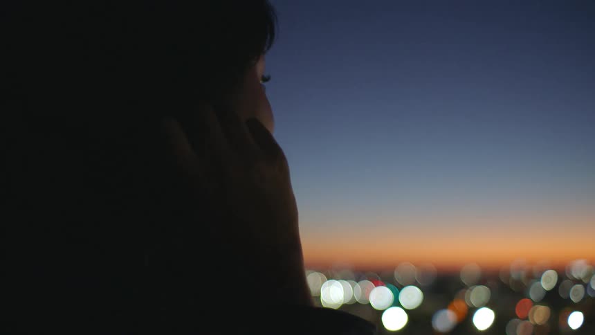 A silhouetted person in side profile gazes out over a softly blurred cityscape at dusk where warm orange and cool blue gradients blend across the sky and distant lights