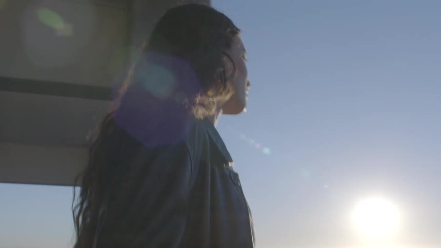 A woman with long dark hair stands outdoors during sunset looking out from a concrete structure sunlight creates a bright lens flare across the frame as the camera circles her profile