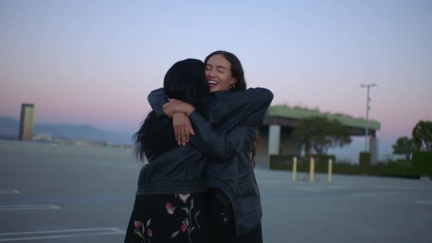 Two young women hug each other on the roof of a parking garage at midday both wearing jackets
