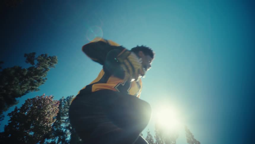 A football player wearing a yellow jersey with the number 17 leaps over a lowangle camerathe scene features a bright sun and tall evergreen trees set against a clear blue sky