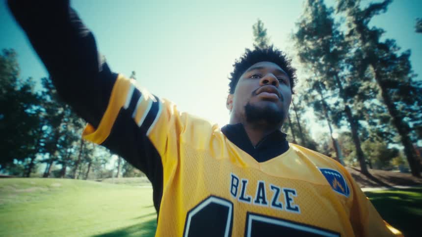 An american football player in a yellow and black blaze jersey prepares to throw a football outdoors on a sunny day the camera captures a lowangle shot as he winds up and releases the ball against a backdrop of tall trees and bright sunlight