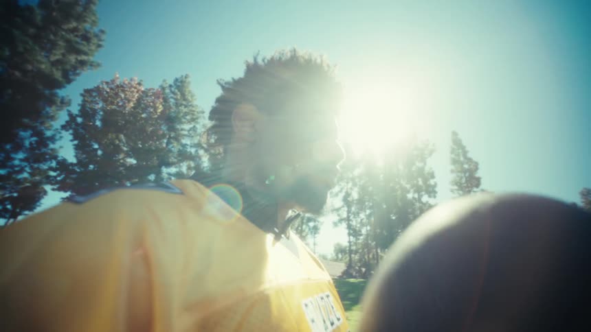 A young black man in a yellow football jersey and gloves stands on a sunlit grassy field holding a black and yellow football with tall trees and a clear blue sky stretching out behind him