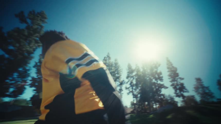 A young football player sprints across a sundrenched field carrying a ball and ducking past the camera before running off toward the distant trees