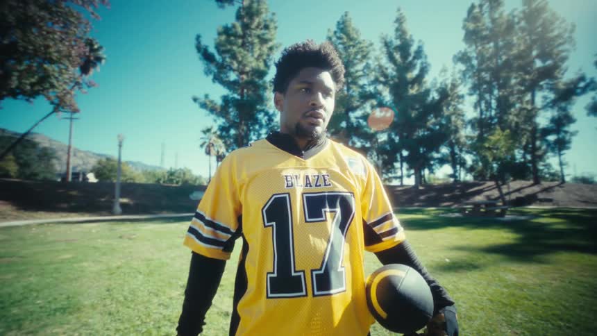 A man wearing a yellow football jersey with the number seventeen and black pants stands on a green field holding a football while looking toward the camera on a bright sunny day with trees in the background