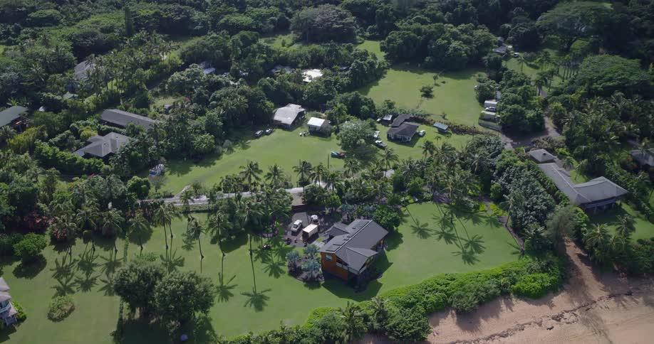 An aerial view of a lush tropical coastline shows a small cluster of houses and manicured lawns nestled among dense green jungle at the base of steep forested hills with a narrow sandy beach and gentle waves