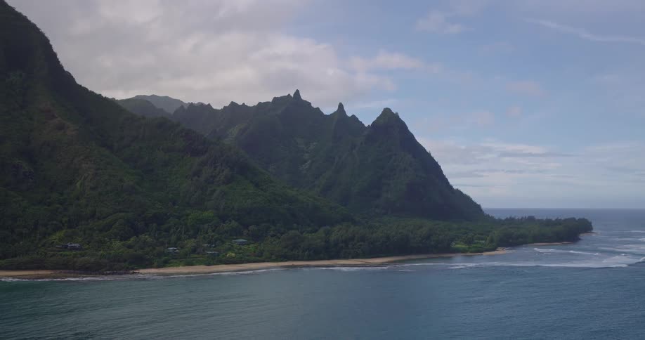Green mountains with sharp peaks border calm beach under partly cloudy sky