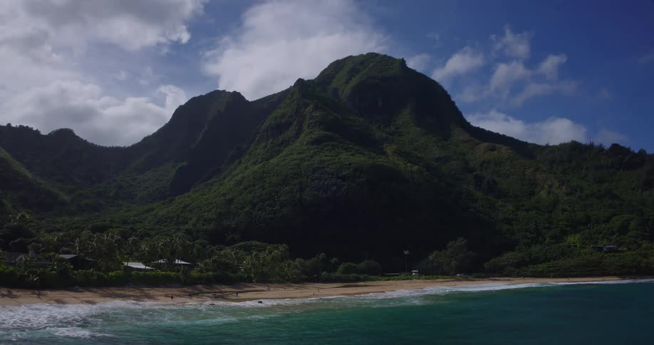 Forested mountains overlook houses and trees along the coast as waves roll in to shore and over the beach