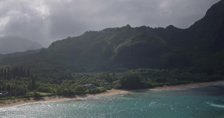 Forested mountains overlook the water with waves rolling over the beach and houses among trees and plants on the coast