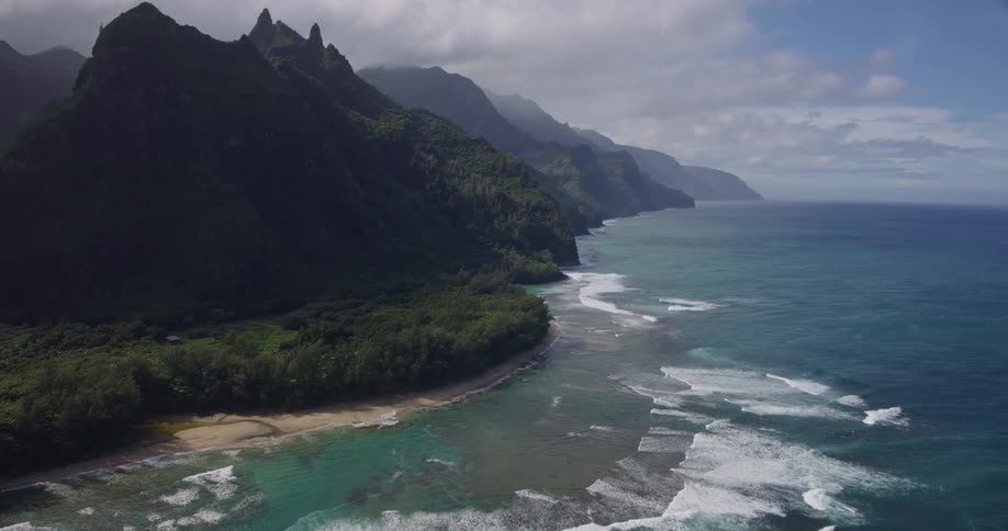 The rugged highcontrast silhouette of the mountain peaks meeting the soft overcast sky the winding shoreline where the turquoise ocean waves crash against the sandy beach and dense green forest