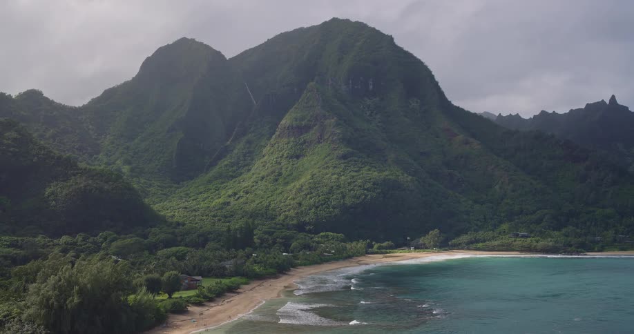 An aerial shot captures a lush mountainous coastline where steep green peaks meet a crescentshaped sandy beach and turquoise oceanthin waterfalls cascade down the dark rock faces as gentle waves wash over the shoreline under a hazy overcast sky
