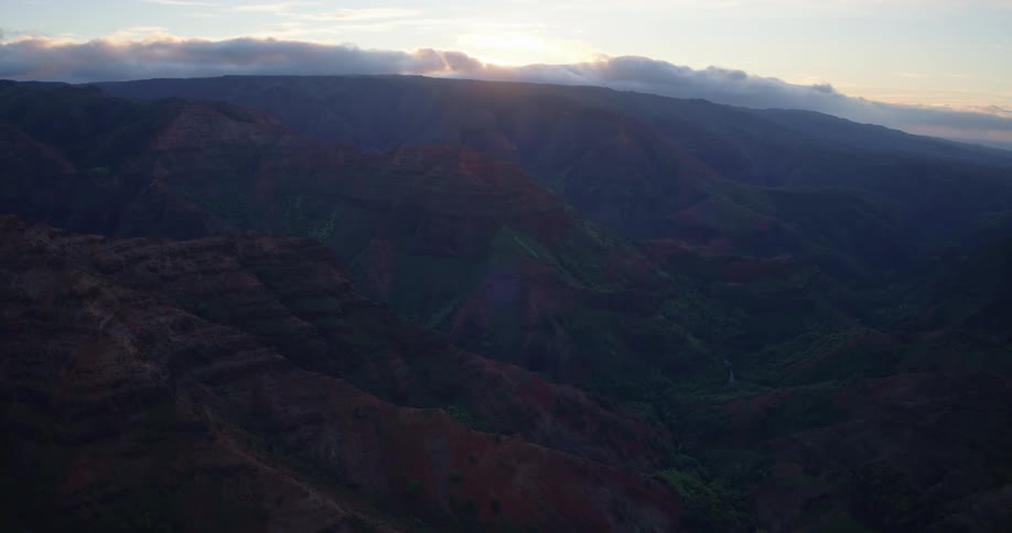 The sun sets behind a layer of clouds casting soft light over the vast rugged ridges and deep shadows of a wide mountain canyon