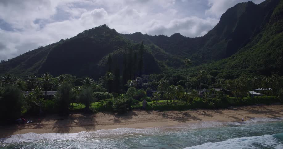 An aerial view captures a secluded sandy beach with palm trees and houses bordering lush dark green dramatic mountains under a partially cloudy sky