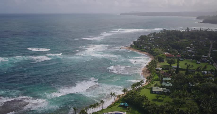 An aerial view of a tropical coastline shows turquoise waves breaking onto a narrow sandy beach lined with palm trees while lush green residential properties and winding roads stretch inland beneath a hazy overcast sky