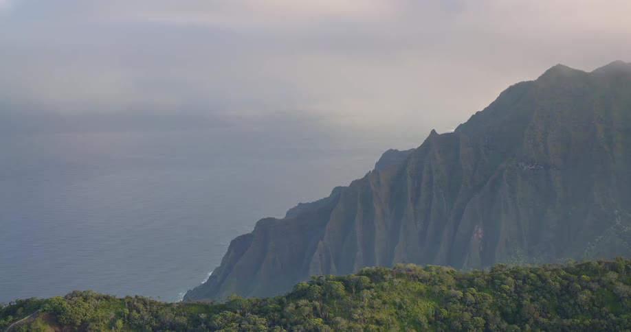 An aerial camera glides past a massive jagged mountain ridge with deep vertical grooves that descends into a misty sea under an overcast hazy sky