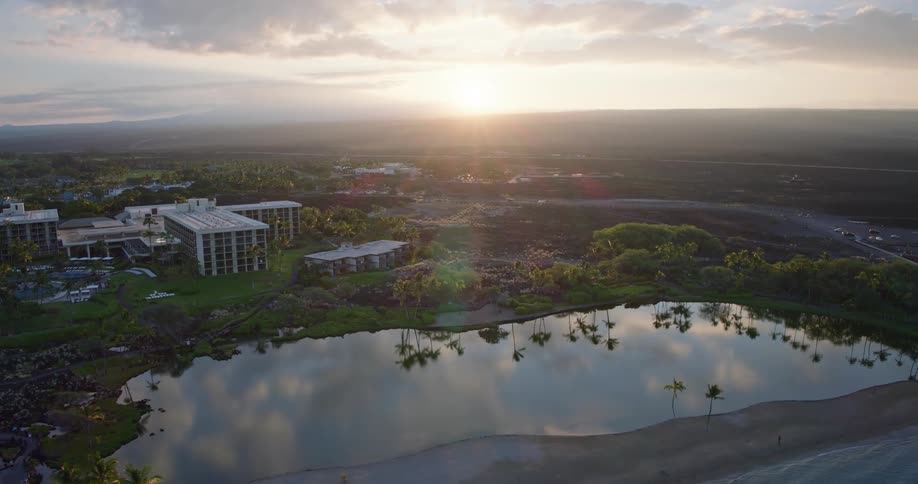 An aerial shot captures a scenic coastal resort with a calm lagoon palm trees and hotel buildings all set against a vast dark volcanic landscape under a bright sunset