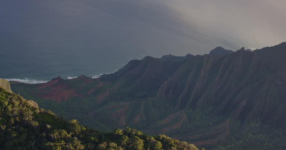 A high angle view captures steep green mountain ridges and red soil cliffs sloping down toward the dark blue ocean under a hazy sunset sky