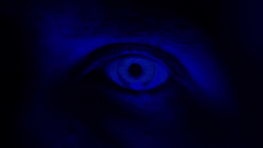 A closeup of a human eye blinking bathed in deep blue light shows the iris and pupil glowing eerily in the darkness creating a dramatic almost otherworldly atmosphere