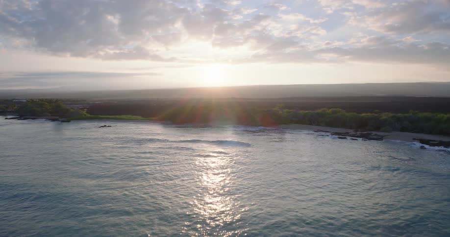 An aerial drone shot glides over turquoise ocean waves toward a rocky coastline at sunset warm sunlight reflects off the waters surface as the camera reveals a lush green shoreline bordering a dark lava field