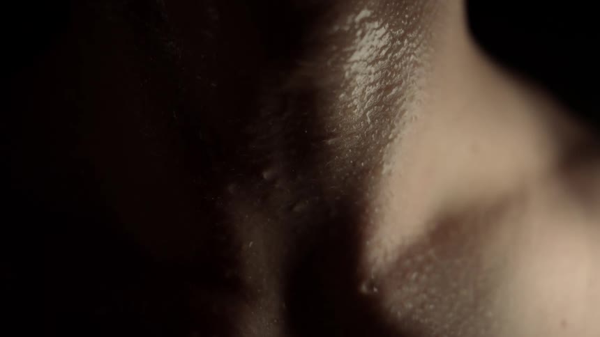 A close view of a man with beads of sweat on his neck and chest in a dark room with low light creating a dramatic atmosphere