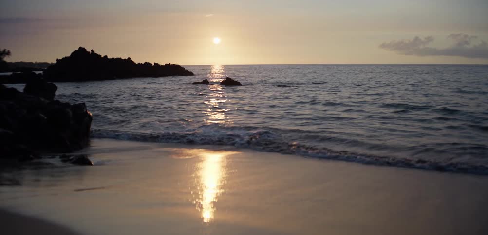The sun sets low over a calm ocean casting a bright golden reflection across the water and onto the wet sand of a shoreline gentle waves lap against the beach near dark volcanic rock formations that sit along the edge of the water
