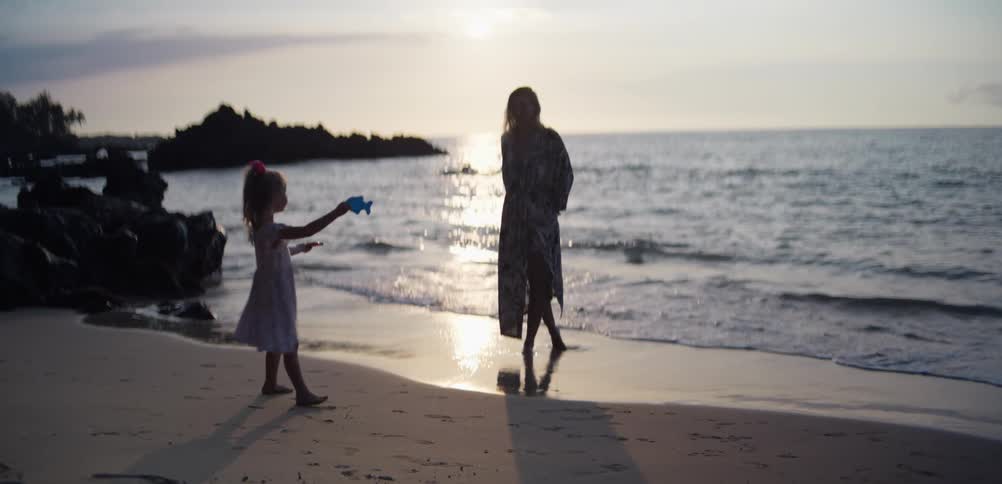 Woman walks along wet sand while a child runs on the beach at sunset