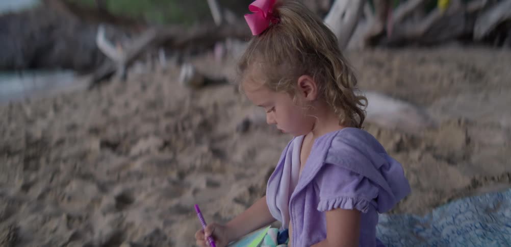 Young girl sitting on beach sand at midday writing in a book with a pen under the bright sun