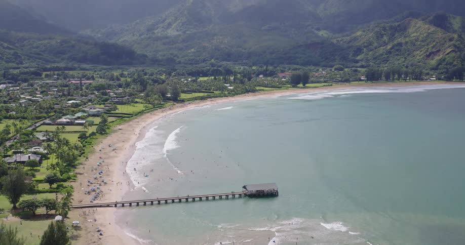 A wide view from above shows a long wooden pier extending into the calm turquoise waters of a crescent shaped bay lined with a sandy beach and lush green hills under a bright sky