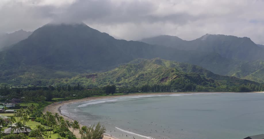 An aerial view glides across a crescentshaped bay and beach toward a small coastal town with a long wooden pier situated at the base of steep mistcovered green mountains