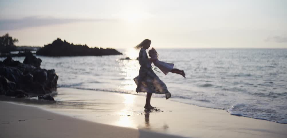 A woman wearing a patterned long dress spins a young girl in a circle on a sandy beach during sunset with the ocean waves and dark rocks in the background