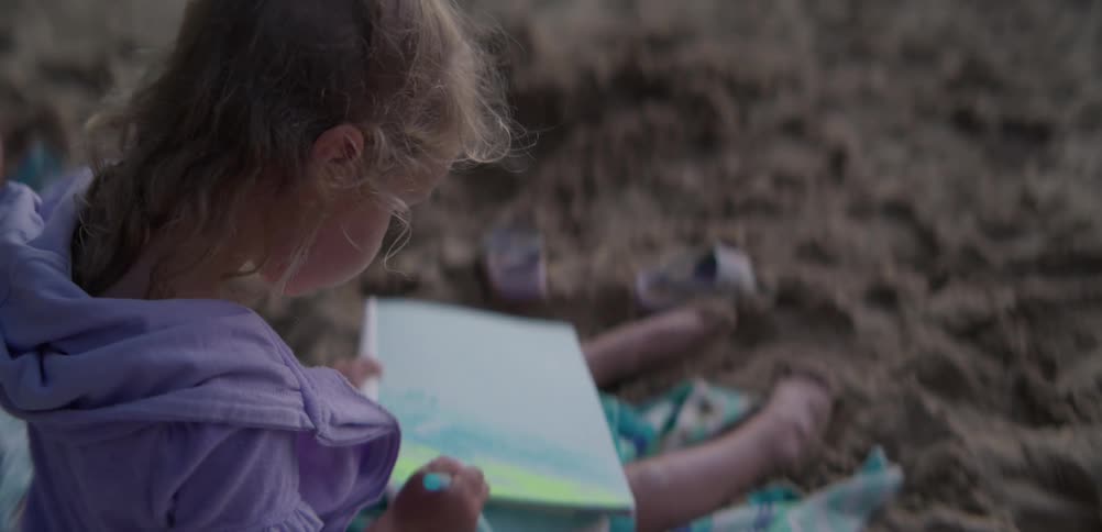 A young girl wearing a purple shirt sits on a striped towel in the sand and uses a blue crayon to draw in a sketchbook on a beach during the evening