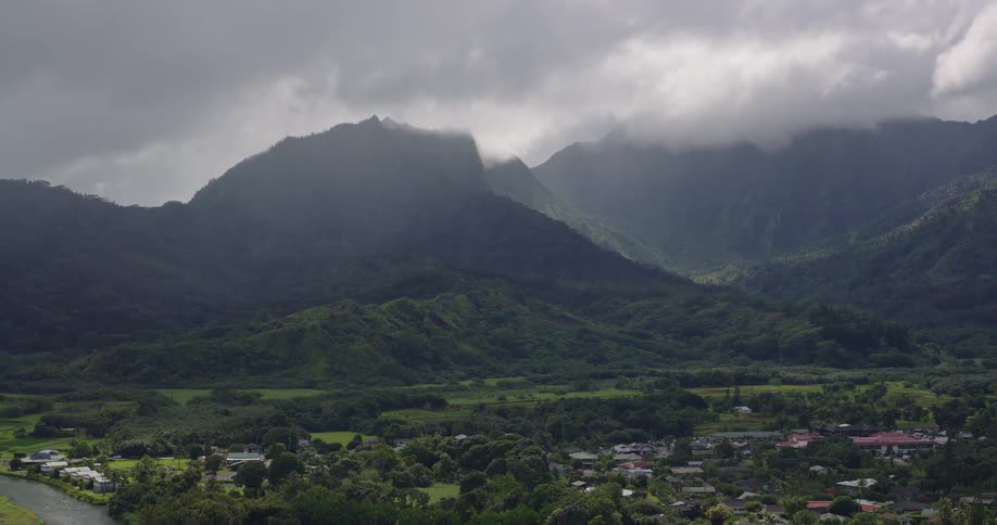 This video features a highaltitude aerial view of a lush mountainous landscape under a heavy overcast sky the camera slowly zooms out to reveal a small town and a winding river nestled at the foot of the misty green peaks