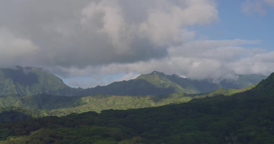 This scenic wide shot captures a lush green mountain range under a dynamic sky filled with large drifting white and gray clouds as the clouds move they cast shifting shadows across the verdant slopes and the dense forest canopy in the foreground