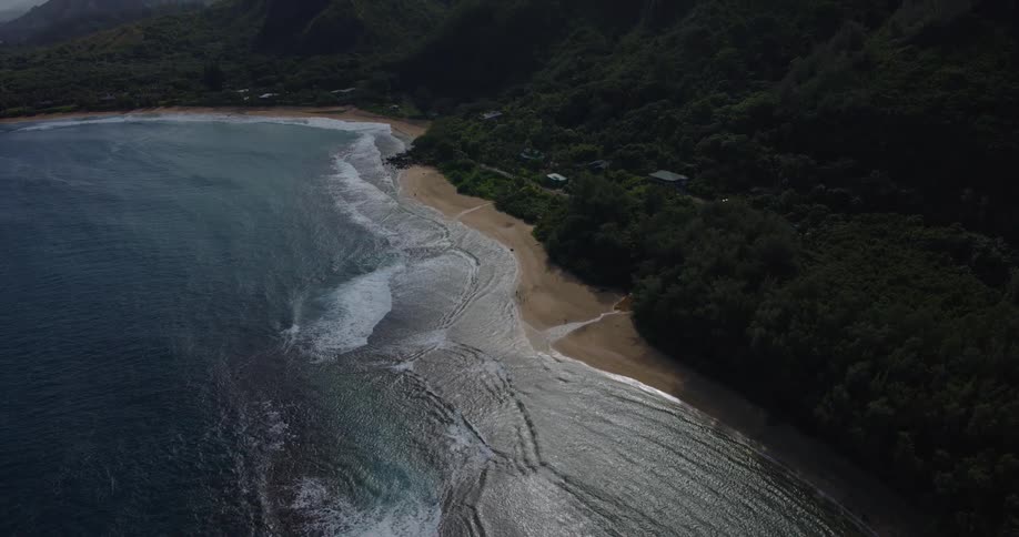 High angle view of a tropical coastline where white waves crash onto a sandy beach bordered by lush green forests and steep mountains under a blue sky with soft clouds