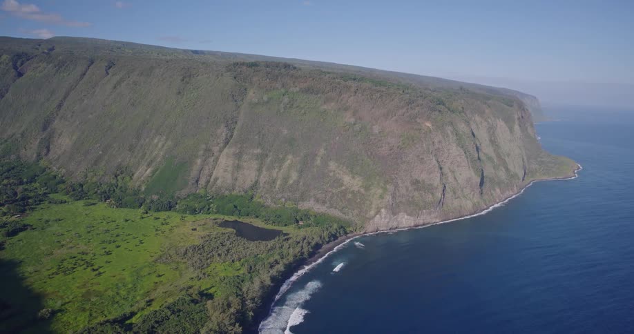 An aerial view reveals a dramatic lush green cliffside plunging directly into the deep blue oceanwaves crash against a narrow dark shoreline at the base of the massive flattopped ridge