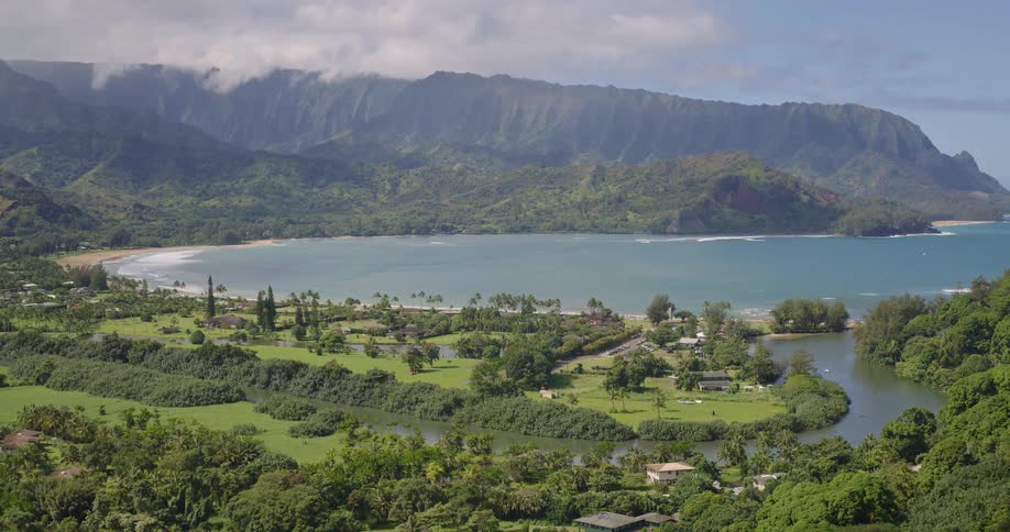 A wide aerial view shows a lush green valley meeting a crescentshaped bay under dramatic cloudcapped mountains a winding river flows through the tropical landscape as sunlight shifts across the verdant hills and calm blue water