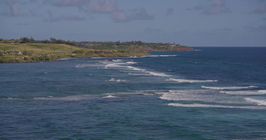 A highangle view captures long white waves rolling across a deep blue ocean toward a green coastline dotted with small houses and utility poles under a clear sky