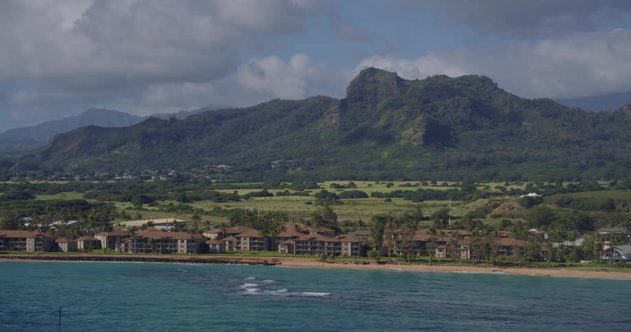 A row of multistory coastal resorts sits along a sandy beach nestled between turquoise ocean waters and lush green fieldsbehind the coastal development a jagged treecovered mountain peak rises sharply into a sky filled with soft white clouds
