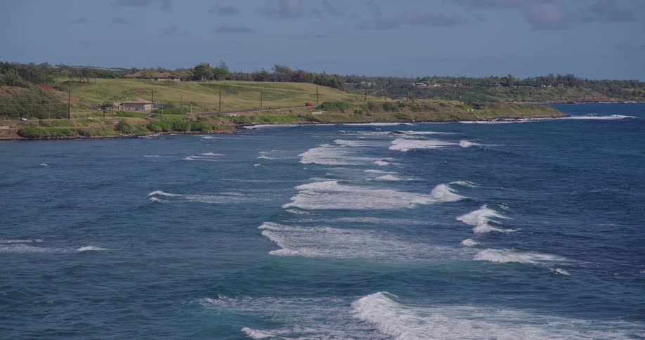 Waves with white foam roll in parallel lines towards a green hilly coastline with a road and a few houses under a partly cloudy sky