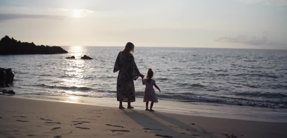 A woman and a young child stand handinhand on a sandy beach as the sun sets over the ocean their silhouettes are framed by the shimmering water and gentle waves washing onto the shore in the soft evening light