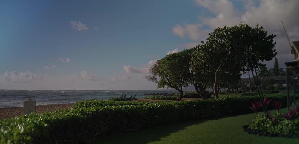 A manicured oceanfront lawn bordered by neatly trimmed hedges and tropical trees overlooks a sandy beach and calm waves under a blue sky dotted with soft clouds with a house and landscaped garden visible on the right side