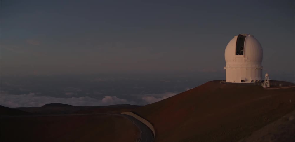 A large white astronomical observatory sits atop a barren reddish mountain ridge overlooking a thick layer of clouds a winding road snakes along the dark hillside leading toward the dome structure under a dim twilight sky