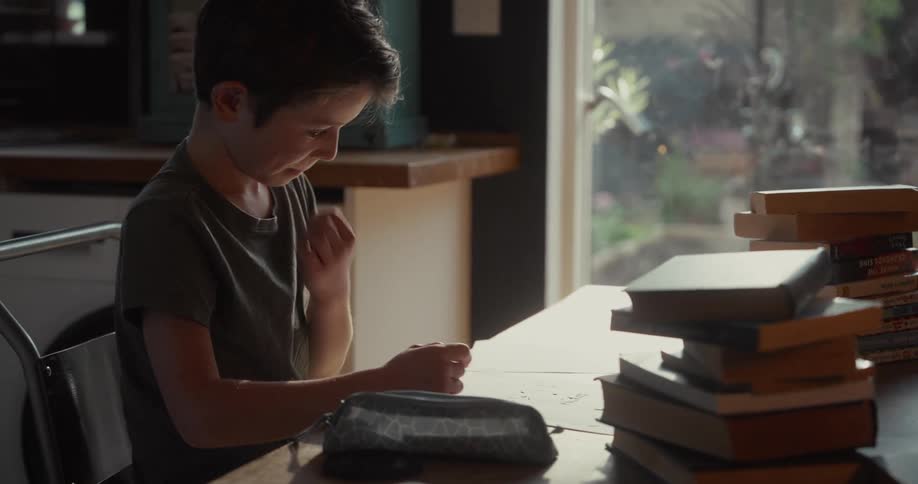 A young boy sits at a wooden table cluttered with stacks of books while focused on writing or drawinghe pauses his work to rub his eyes and face appearing tired or frustrated by his task